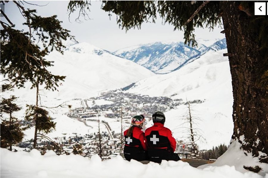Two ski patrol members in red uniforms sitting on a snowy mountainside overlooking the town of Ketchum